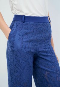 Pantalones azules bordados con un patrón texturizado, que cuentan con una cinturilla lisa y bolsillos laterales. La tela ligera muestra detalles de diseño intrincados.