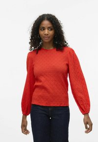 Rotes, strukturiertes Langarmoberteil mit rundem Halsausschnitt und gerafften Manschetten, kombiniert mit dunklen Denim-Jeans. Der Stoff weist ein dezentes Muster auf.