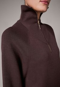 Fioletowy sweter dzianinowy z wysokim kołnierzem, wyposażony w zamek błyskawiczny i subtelne przeszycia na ramionach. Miękka tekstura i luźny krój.