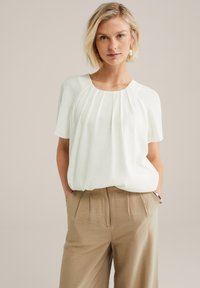 Blouse blanche plissée à manches courtes et col rond, associée à un pantalon beige texturé, avec poches latérales et coupe décontractée.