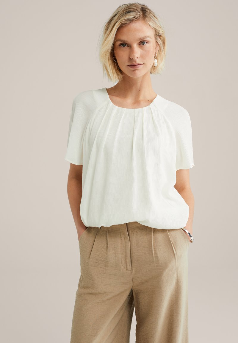 Blouse blanche plissée à manches courtes et col rond, associée à un pantalon beige texturé, avec poches latérales et coupe décontractée.