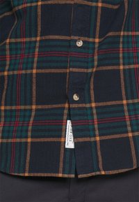 Camisa de franela con base azul marino y patrón de cuadros en verde, rojo y dorado, cierre de botones y una pequeña etiqueta de la marca cerca del dobladillo.