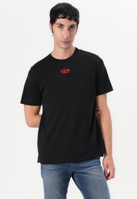 Zwarte katoenen T-shirt met ronde hals, korte mouwen en een rood logo op de borst. G gedragen met lichtblauwe denim jeans.