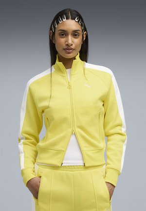 Vrouw met steil haar gestyled met meerdere witte haarspelden, gekleed in een geel-witte jas met rits en bijpassende broek, met handen in de zakken.