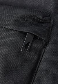 Primer plano de una bolsa de tela negra con un bolsillo con cremallera y un clip de plástico negro. La textura es suave con detalles de costura visibles.