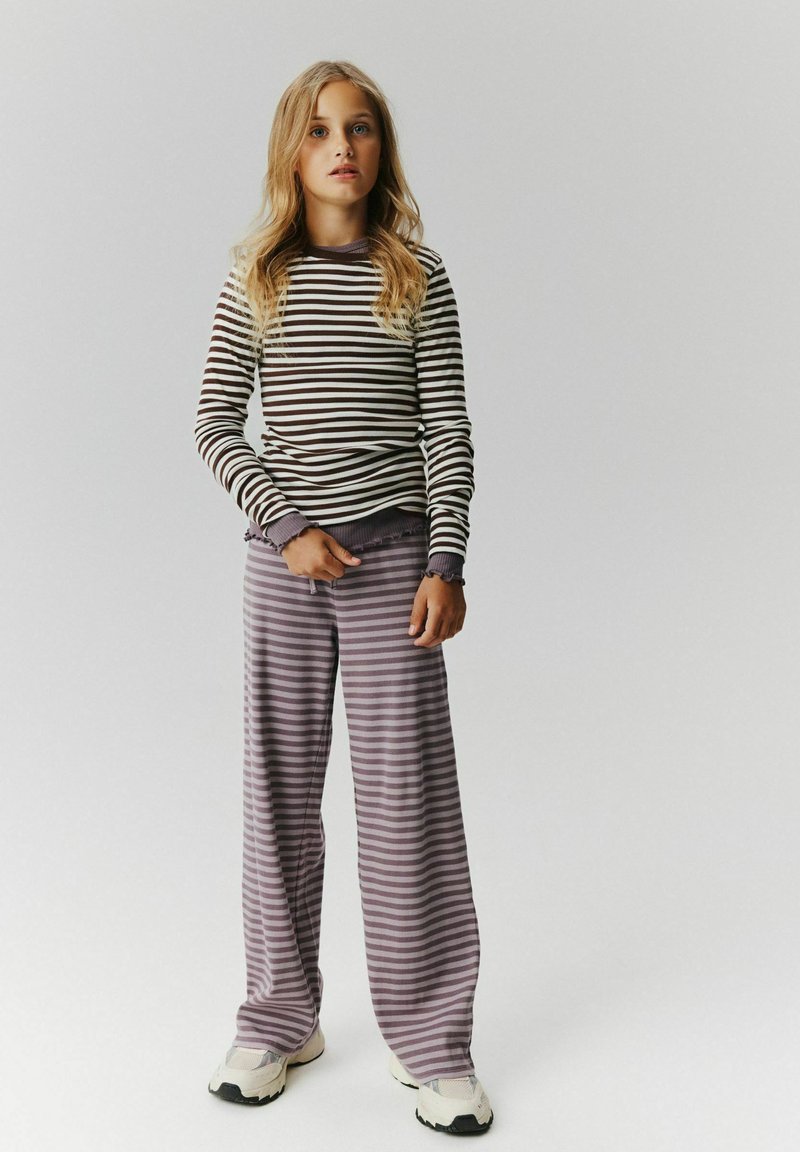 Jeune fille aux longs cheveux blonds portant un t-shirt à manches longues rayé et un pantalon large rayé avec des baskets blanches.