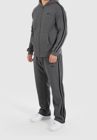 Grau zip-up Hoodie und passende Hose aus weichem Material, mit schwarzen Seitenstreifen und einem Logo auf der Brust. Weiße Sneaker runden den Look ab.