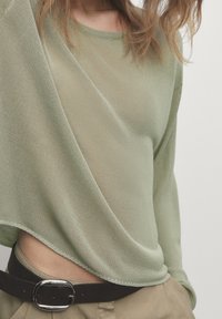 Pull ample en maille vert clair avec un tissage texturé. Manches longues, encolure large et ourlet irrégulier, associé à une ceinture marron.
