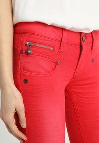 Vaqueros de mezclilla rojos con un bolsillo de cremallera metálica, detalles de costura y un pequeño logo en el bolsillo frontal. Textura lisa y diseño ajustado.