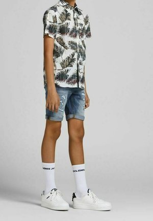 Lyhythihainen, valkoinen napillinen paita, jossa on vihreä ja musta palmunlehtikuvio, yhdessä kuluneiden farkkushortsien ja valkoisten tennareiden kanssa.