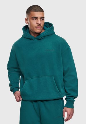 Mężczyzna w miętowym kapturze Dropsize i dopasowanych spodniach dresowych, patrzący na bok z neutralnym wyrazem twarzy na jasnym tle.