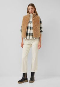 Gilet di pelliccia tan sopra una camicia a quadri con tonalità verdi e beige, pantaloni crema con orlo sfrangiato e stivali neri con suola spessa. Sfondo neutro.