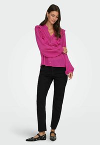 Fuchsia Bluse mit Rüschendetails, langen Ärmeln und Knopfleiste, kombiniert mit schwarzen Slim-Fit-Jeans und besetzen schwarzen Ballerinas.