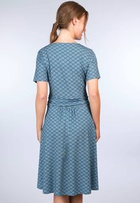 Eine Rückansicht eines Kurzarmkleides aus türkisfarbener Stoff mit einem Blattmuster, tailliert und mit ausgestellt gesäumtem Rock.