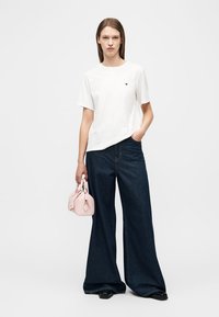 Γυναίκα που φοράει λευκό πουκάμισο με κοντά μανίκια και σκούρα τζιν με φαρδιά μπατζάκια, κρατώντας μια μικρή ροζ τσάντα, στέκεται μπροστά σε ένα απλό φόντο.