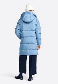 Veste doudoune bleue avec une capuche, présentant un design matelassé et des zips sur les côtés, associée à un pantalon sombre et des chaussures noires.