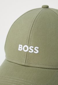 Olivengrønn caps laget av strukturert stoff har en hvit brodert "BOSS" logo foran. Kurvet skjerm og sydd detaljer er synlige.