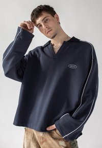 Marinblå sweatshirt med V-hals, vit kantning och framlogotyp. Har långa, breda ärmar och en avslappnad passform. Texturerat tyg.