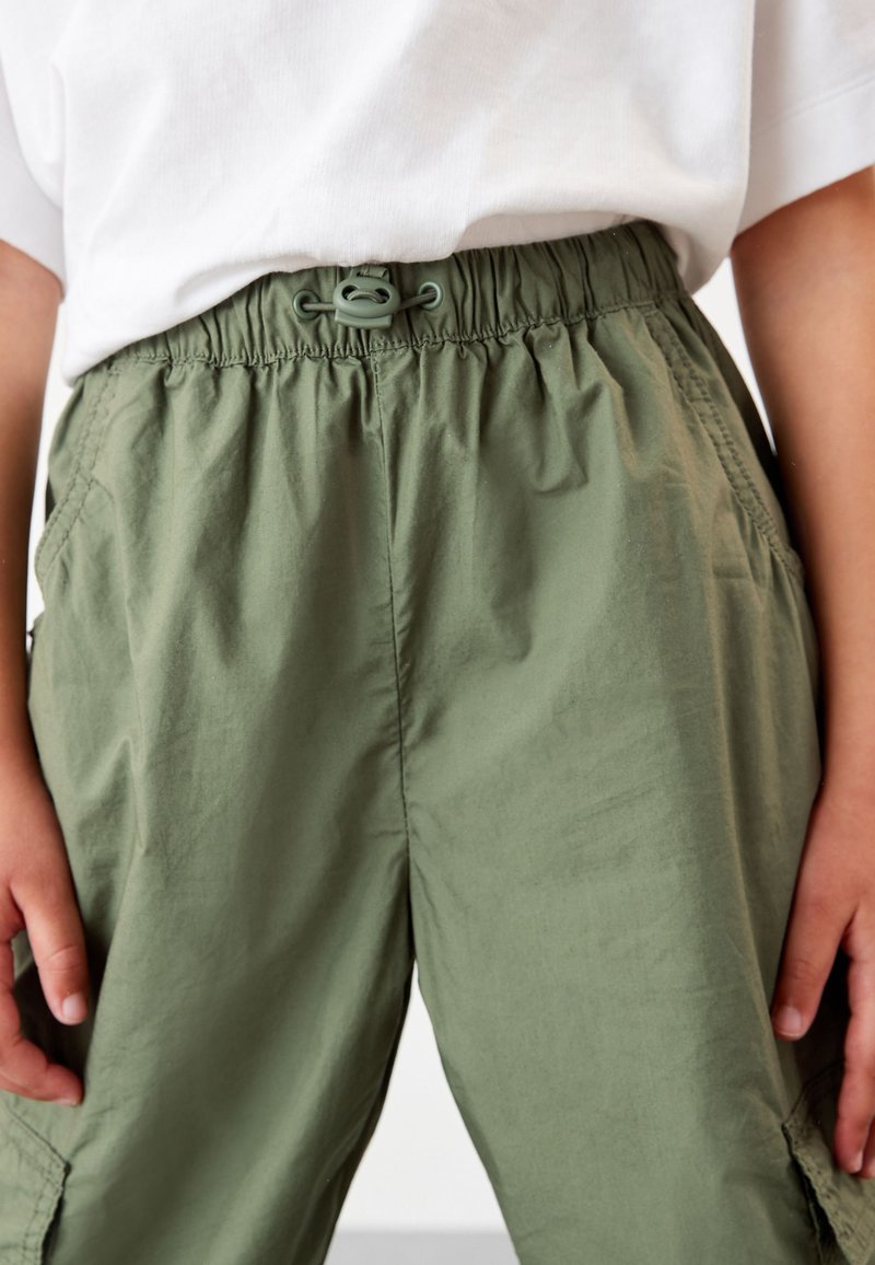Enfant portant un pantalon cargo vert olive avec une taille élastique et un cordon, associé à un t-shirt blanc à manches courtes.