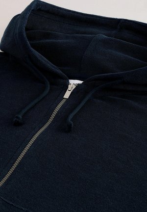 Navy plava zip-up dukserica izrađena od mekanog materijala, s kapuljačom, srebrnim patentnim zatvaračem i vezicama. Ima teksturirani izgled s ležernim dizajnom.