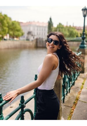 Bílý tílko, černé kalhoty, dlouhé vlnité vlasy a černé nadrozměrné sluneční brýle. Stojí u zeleného zábradlí poblíž vody, s rozmazanými budovami v pozadí.