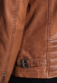 Giacca di pelle marrone con una texture liscia, dotata di una cerniera laterale, un dettaglio a tracolla con fibbia e accenti cuciti sul retro.