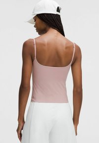 Light pink sleeveless top with thin straps, smooth texture, fitted design, and a subtle logo at the hem. Worn with white pants.