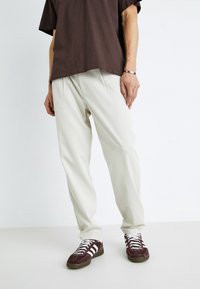 Pantalons crème clair à coupe décontractée, avec un front plié, associés à une chemise à manches courtes marron foncé et des baskets bordeaux avec des rayures blanches.