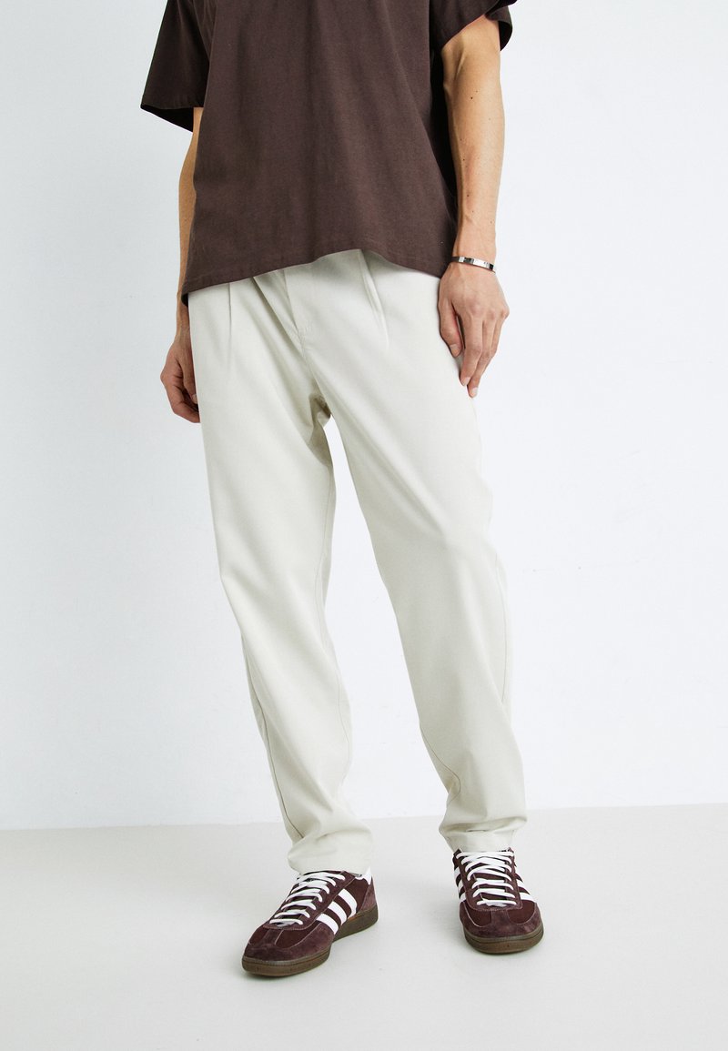 Pantalons crème clair à coupe décontractée, avec un front plié, associés à une chemise à manches courtes marron foncé et des baskets bordeaux avec des rayures blanches.
