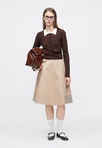 Καφέ πλεκτή ζακέτα με λευκή γιακά, μπεζ φούστα σε Α γραμμή με καρό τελείωμα και καφέ δερμάτινη τσάντα. Λευκές κάλτσες έως το γόνατο και μαύρα παπούτσια.