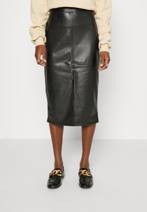 Vrouw draagt een knielange zwarte leren kokerrok, een crèmekleurige top met lange mouwen en zwarte loafers met grote gouden kettingdetails.