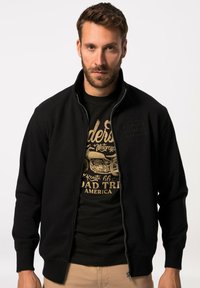 Giacca nera con zip e colletto alto, caratterizzata da un design texture sul petto, indossata sopra una t-shirt nera con grafica e stampa dorata.