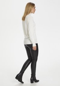 Pulover alb cu guler înalt și mâneci lungi, asortat cu pantaloni negri, mulați, din piele ecologică și botine, evidențiind o siluetă elegantă.