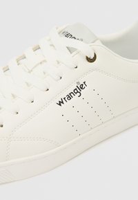 Vita lädersneakers med rund tå, texturerad sula, platta vita snören och varumärkeslogotyp. Har perforeringar för ventilation nära sulan.