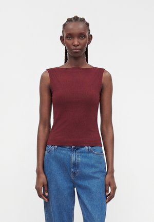 Kvinde med flettet hår iført ærmeløs tætsiddende burgundy top og højtaljede blå jeans, stående mod en ensfarvet lys baggrund.