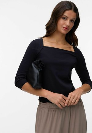 Femme en haut noir à encolure carrée et jupe beige tenant une pochette en cuir noir pliée sous son bras sur un fond blanc uni.