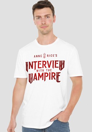 Young man wearing a white T-shirt with red text reading "Anne Rice's Interview with the Vampire" against a plain gray background.