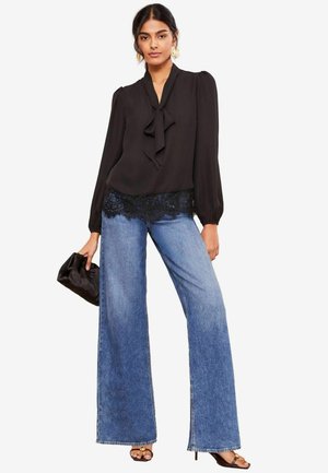 Black blouse with a bow tie at the collar and lace trim. Paired with wide-leg blue denim jeans and a black clutch handbag.