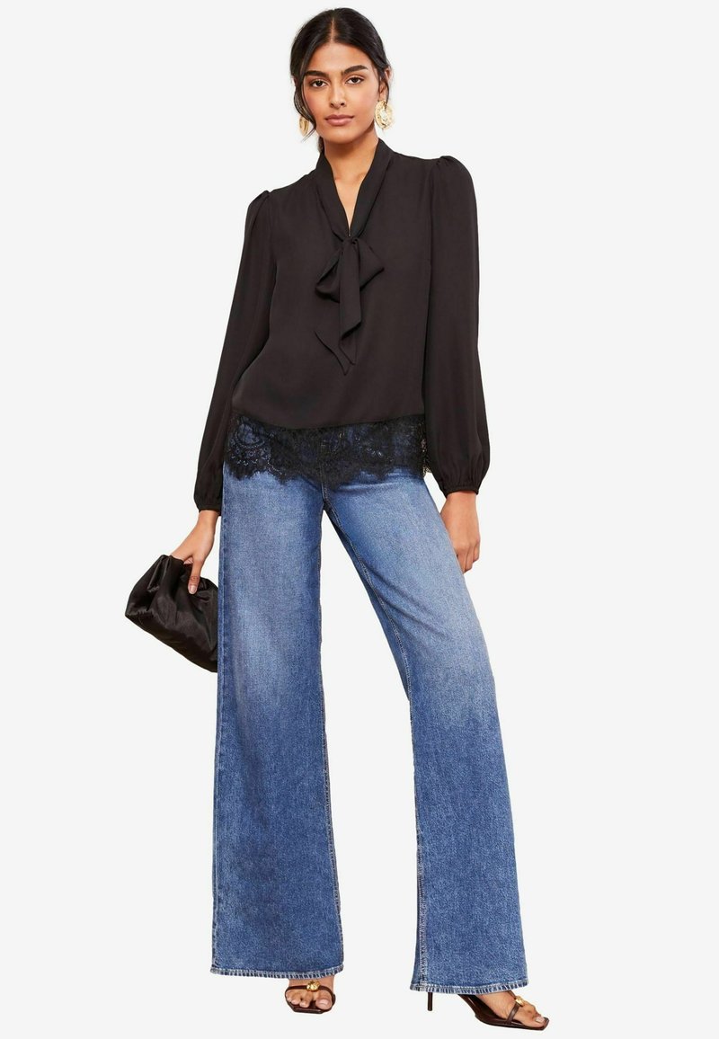 Zwarte blouse met een strik bij de kraag en kantafwerking. Gepaarde met wijde blauwe denim jeans en een zwarte clutch handtas.