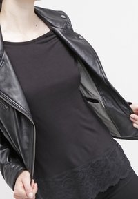 Personen har en svart läderjacka över en svart topp med spetsdetaljer på fållen, vilket visar jackans inre foder och ficka.