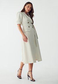 Vestido beige hasta la rodilla con mangas abullonadas, botones de doble botonadura y una cintura ceñida con cinturón. Cuenta con dos bolsillos frontales y un dobladillo recto.