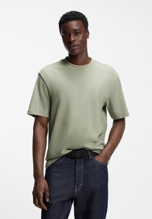 Ung mand iført en lysegrøn kortærmet T-shirt med teksten "HUGO", mørke jeans og sort bælte, stående foran en ensfarvet hvid baggrund.