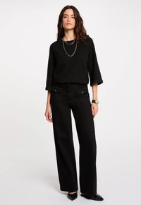 Maglione nero a maglia con maniche a tre quarti, abbinato a pantaloni neri a gamba larga con tasche anteriori. Scarpe nere a punta con borchie.