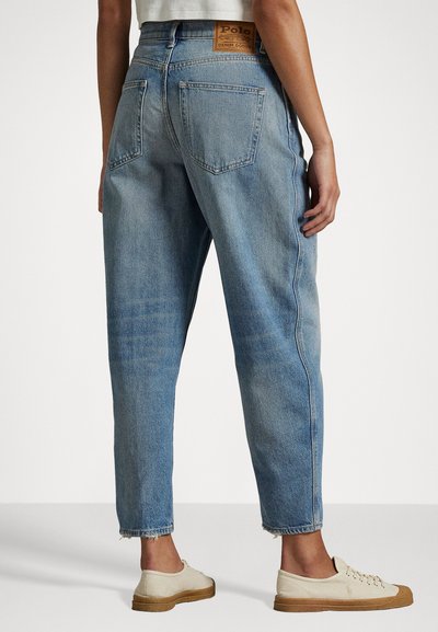 Pessoa vestindo jeans de ganga azul claro com um rótulo Polo na cintura e tênis de tela brancos com solas castanhas.