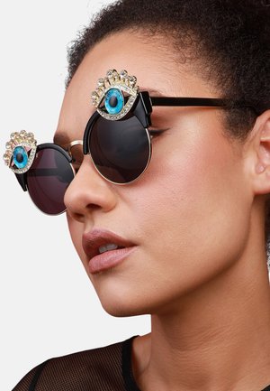 Occhiali da sole rotondi con montatura nera, lenti scure e un design decorativo di occhi jewel sulla parte superiore. Accenti dorati esaltano le caratteristiche.