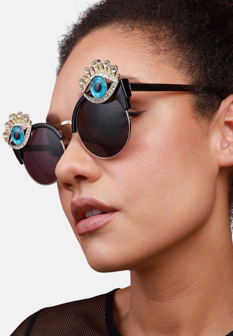 Occhiali da sole rotondi con montatura nera, lenti scure e un design decorativo di occhi jewel sulla parte superiore. Accenti dorati esaltano le caratteristiche.