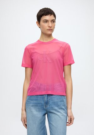 Γυναίκα με κοντά σκούρα μαλλιά φορά διαφανές ροζ t-shirt Calvin Klein πάνω από ροζ τοπ και ανοιχτό μπλε τζιν, στέκεται μπροστά σε απλό λευκό φόντο.