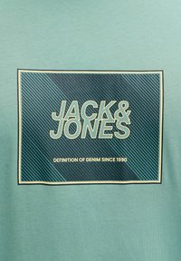Zielona bawełniana koszulka z grafiką w ramce w ciemnym niebieskim kolorze, z białym i złotym napisem "JACK & JONES" oraz "DEFINITION OF DENIM SINCE 1990".