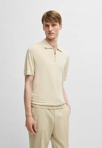 Beige polo skjorta med korta ärmar och dragkedja vid kragen, tillverkad av ett mjukt, lätt tyg. Modellen kombinerar den med texturerade beige byxor.