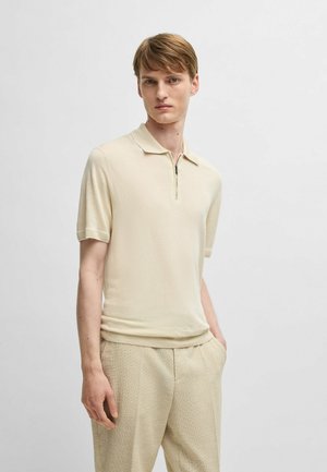 Beige polo t-shirt med korte ærmer og lynlås krave, lavet af et blødt, let stof. Modellen kombinerer det med strukturerede beige bukser.